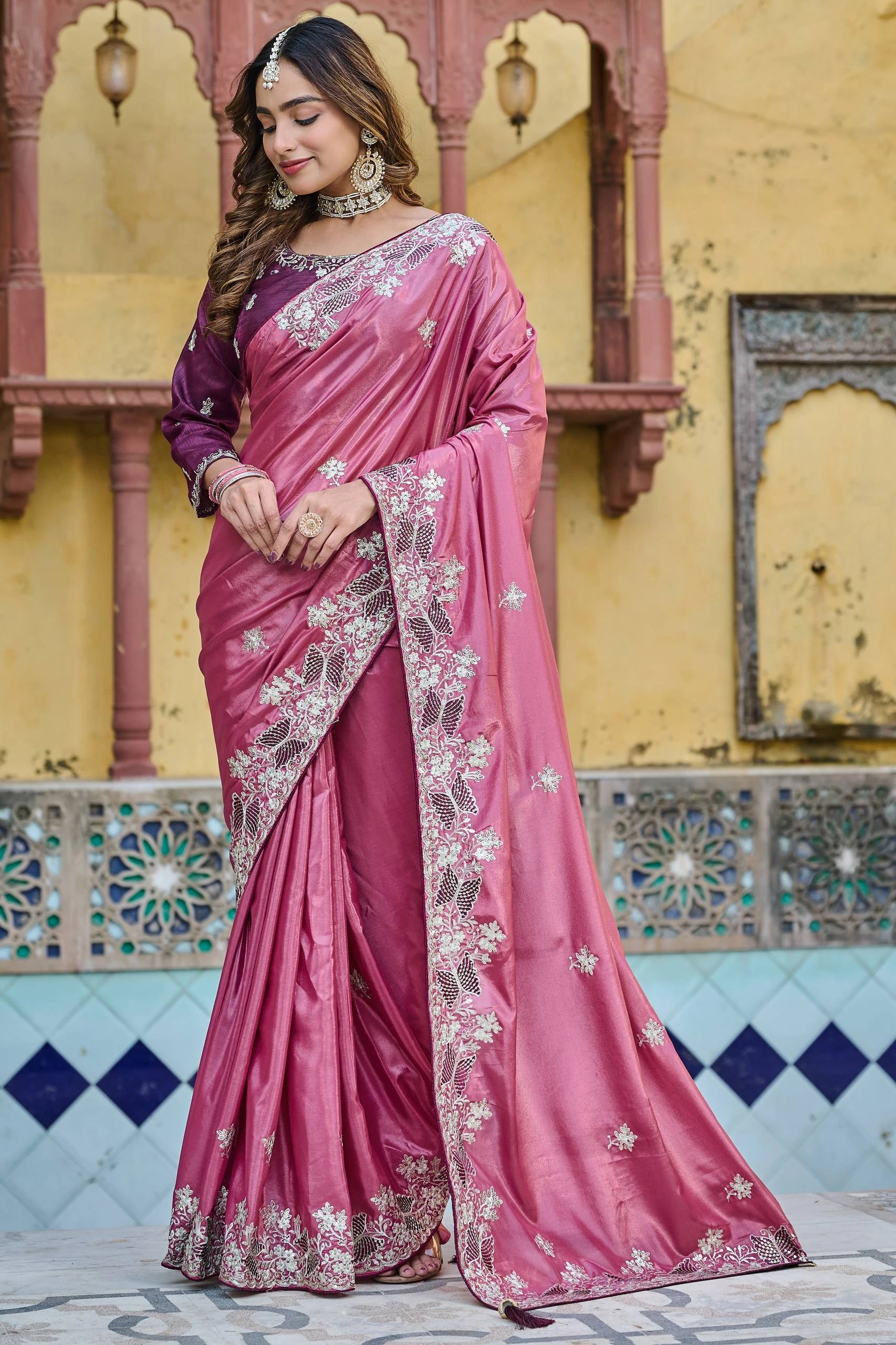 Woman in a pink saree with white embroidery standing in front of a decorative wall.